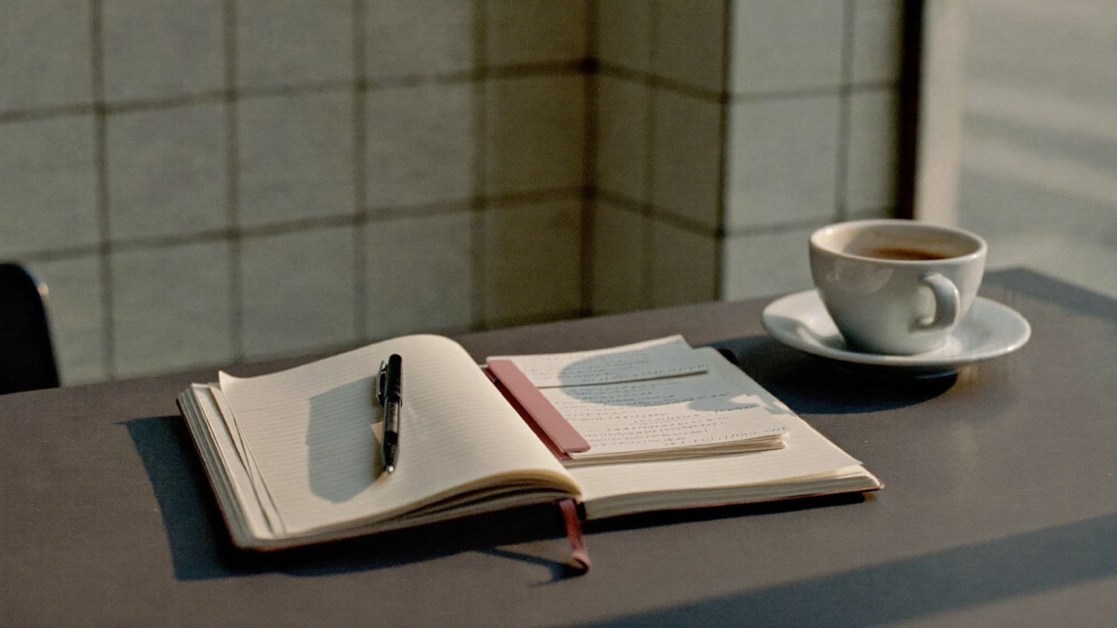 An open ledger notebook with a pen and a stack of receipts held by a dusty pink clip, beside a cup of coffee on a cafe counter