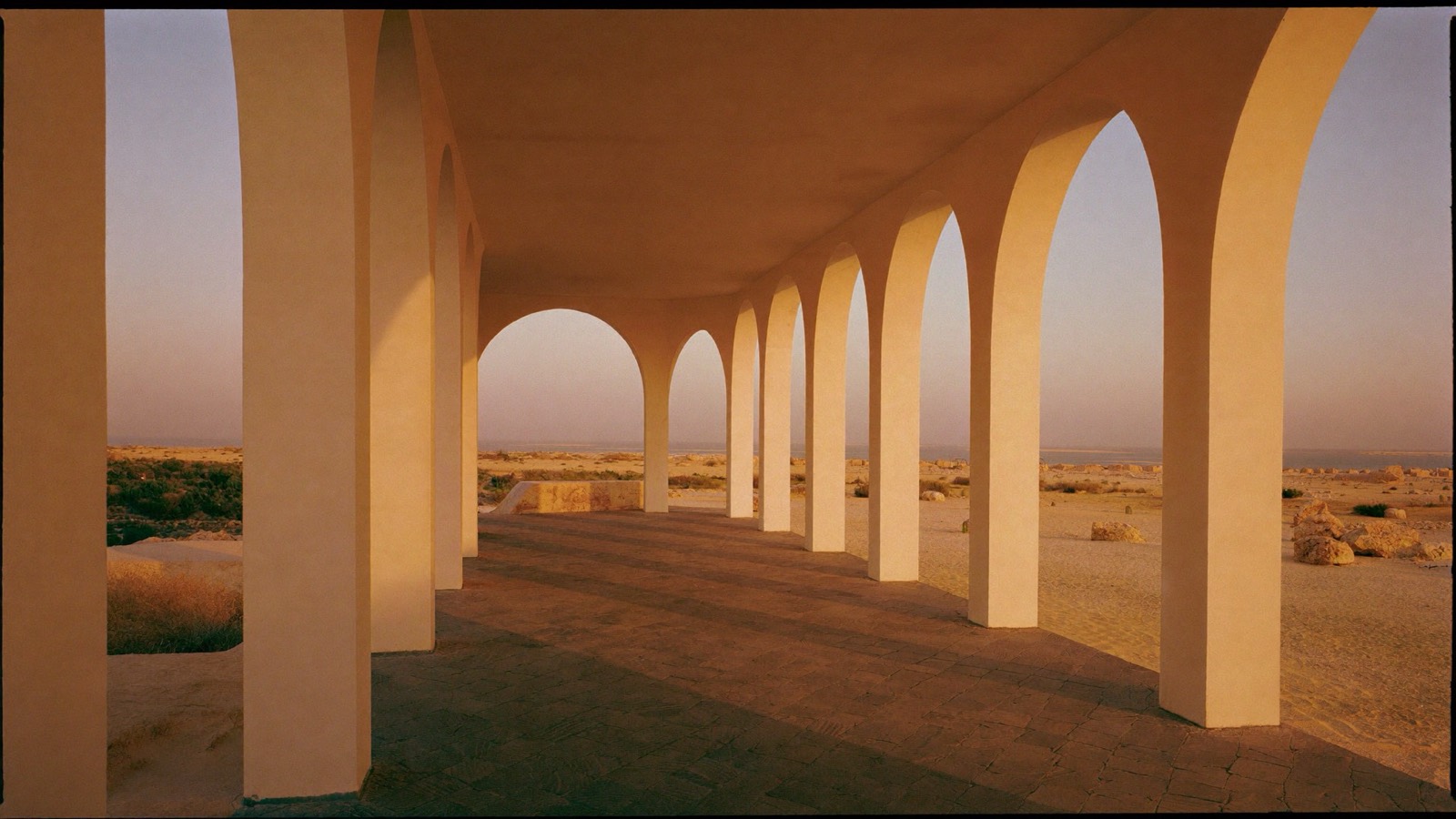 Golden hour light in a colonnade of tall cream stone arches, long amber shafts across the floor