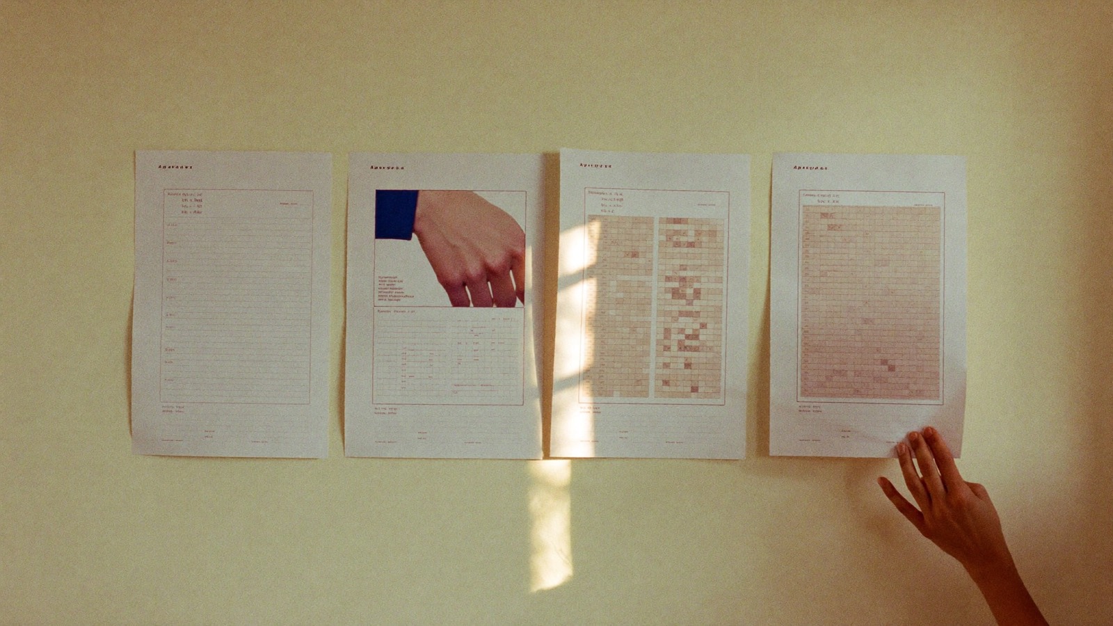 Four printed planning sheets pinned in a row on a warm cream wall, a hand smoothing one corner